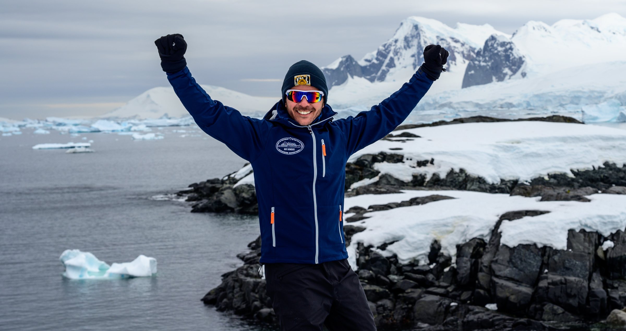 Dan with both arms raised on a rocky point above icebergs and snow-covered mountains on the Antarctic Peninsula