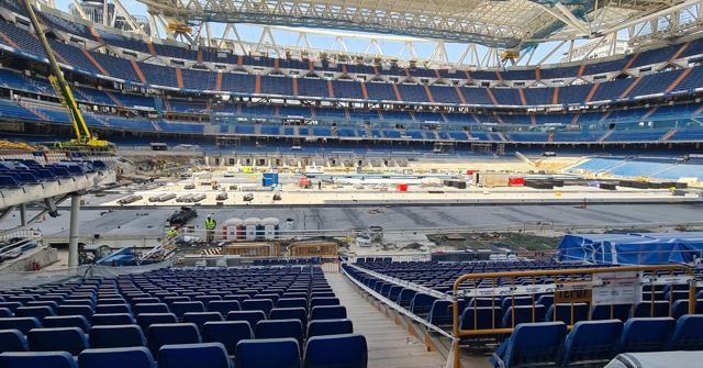 The inside of a large football stadium under renovation, with blue seats and construction work.
