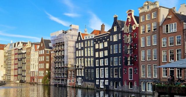 A row of tall, narrow, leaning canal houses reflected in the water under a blue sky.