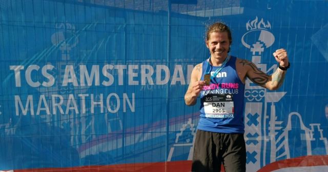 A man in a running vest with a race bib, fist raised, in front of a blue marathon backdrop.