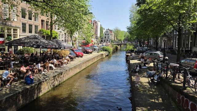 A leafy Amsterdam canal lined with busy cafe terraces and parked bikes on a sunny day.