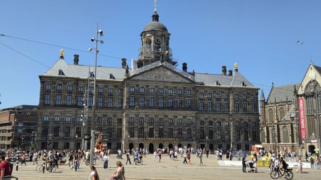 The Royal Palace on Dam Square in Amsterdam with crowds and cyclists in front under a blue sky.