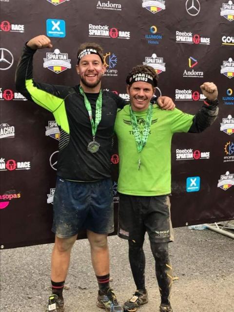 Two muddy men flexing with Spartan race medals in front of a sponsor board.