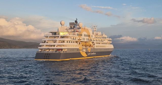 A white and navy expedition cruise ship sailing on calm sea with a headland behind.