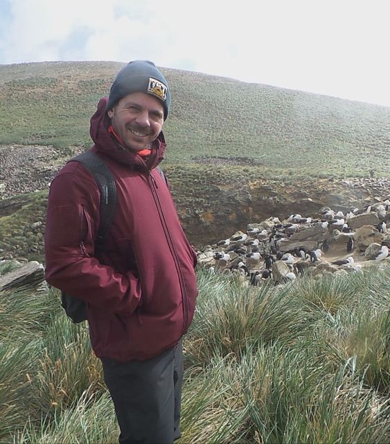 Smiling man in a red coat and beanie standing in tussock grass in front of a penguin colony.