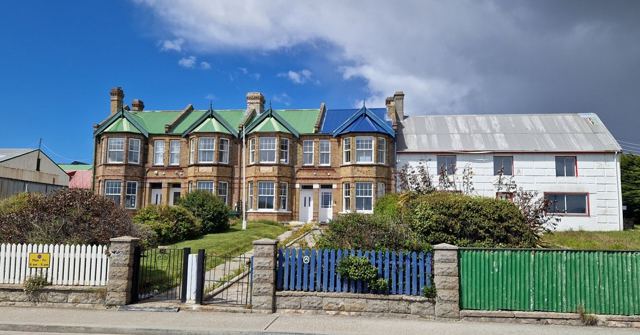 Brick terraced houses with green and blue roofs behind colourful fences under a blue sky.