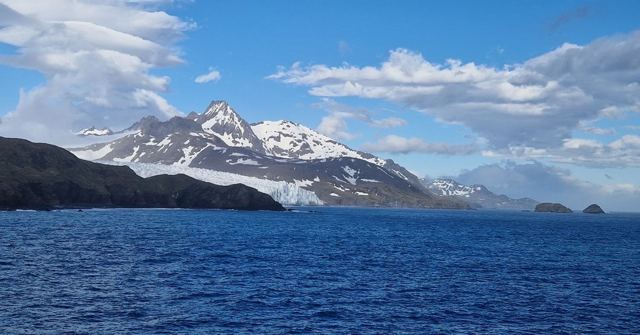 Snowy mountains and glaciers rising above a deep blue sea under a partly cloudy sky.