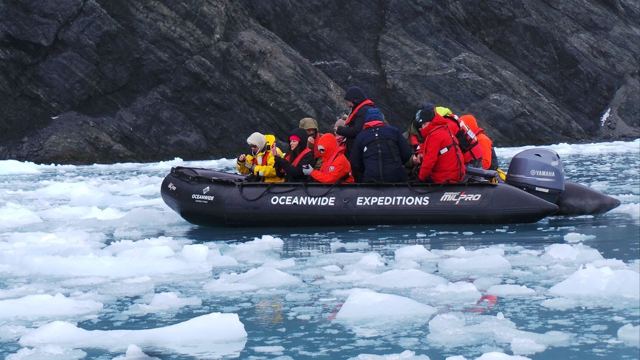 Inflatable boat with passengers in red jackets moving through floating ice by dark cliffs.