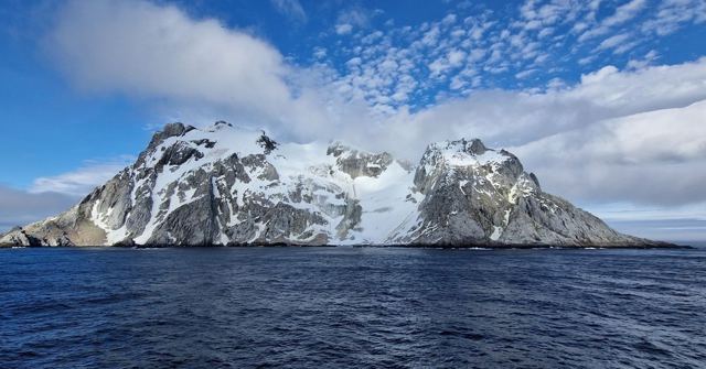Snow-capped rocky island in a dark blue sea under a sky of small clouds.