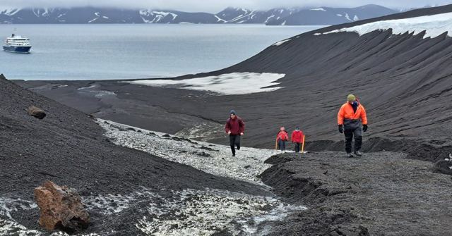 Hikers in red jackets walking up a dark volcanic slope with snow and a ship in the bay.