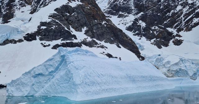 Turquoise-blue iceberg in front of snow-covered rocky mountains.
