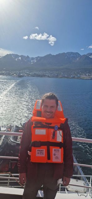 A man in an orange life jacket on a ship's deck with mountains and Ushuaia behind.