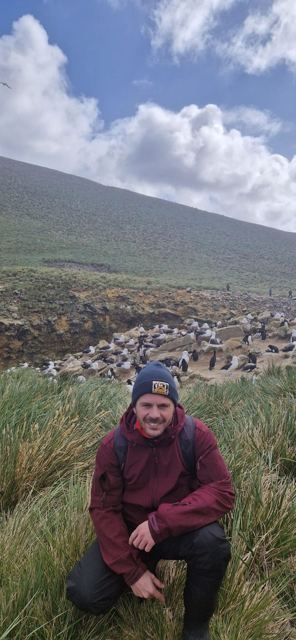 A man in a beanie in front of a hillside colony of nesting seabirds and penguins.