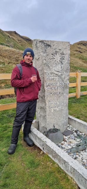 A man in a red jacket holding a small glass beside Shackleton's granite headstone in Grytviken.