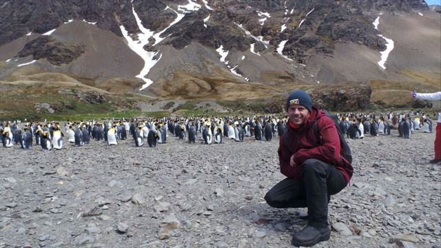 A man crouching on a stony beach in front of a huge colony of king penguins.