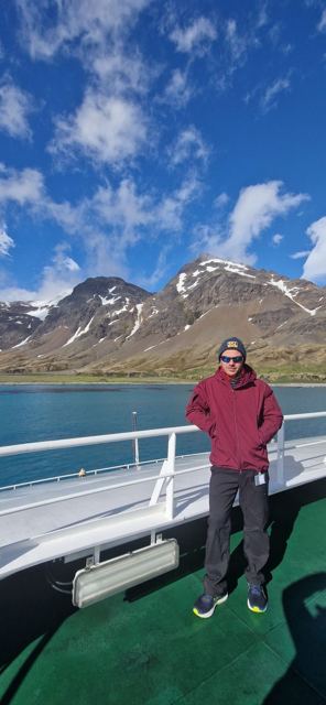 A man in a red jacket on a ship's deck with snow-streaked mountains and a blue bay behind.