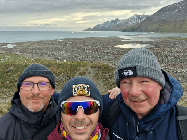 Three men in beanies taking a selfie with a huge penguin colony and mountains behind.
