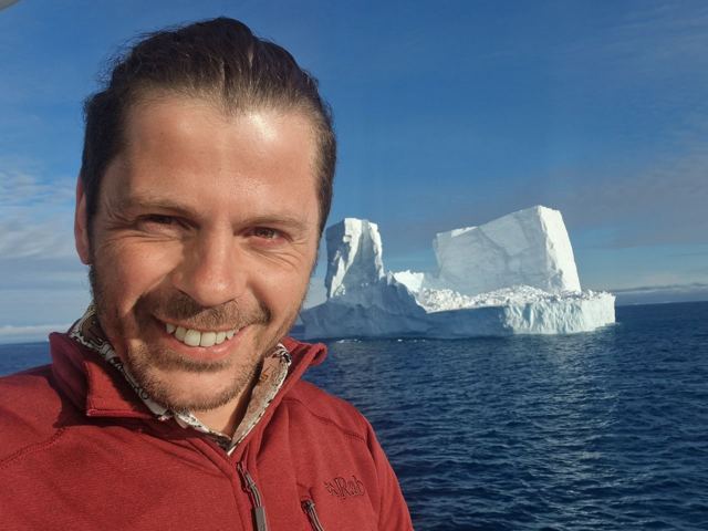 A smiling man taking a selfie on deck with a large flat-topped iceberg in a blue sea behind.