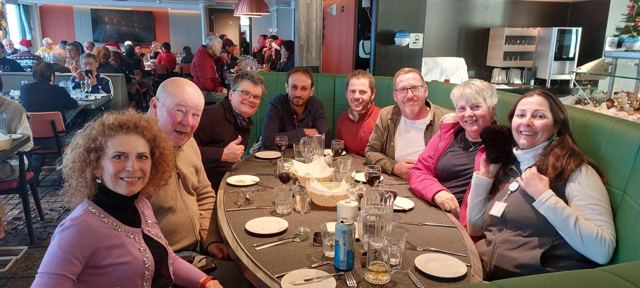 A large group of passengers smiling around a round dining table on a ship.