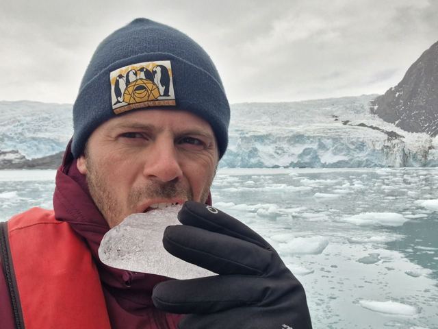 A man in a beanie biting a piece of clear ice in front of a glacier and icy water.