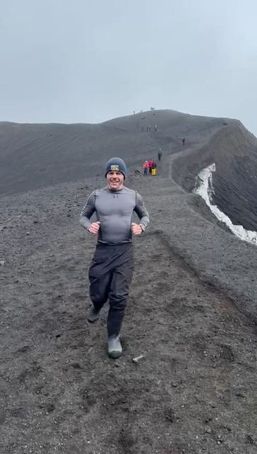 A man in a base layer running along a dark volcanic ridge path with snow nearby.