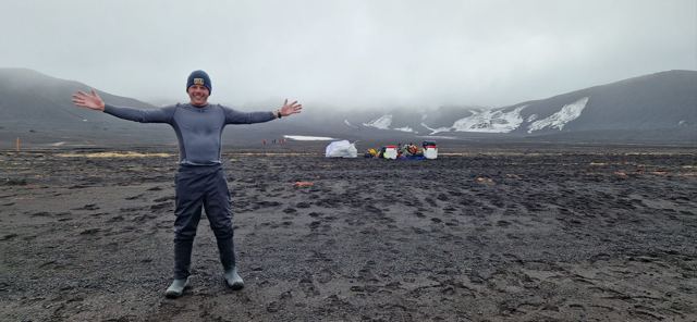 A man with arms outstretched on a dark volcanic beach under grey skies.