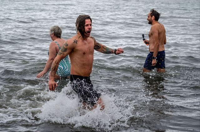 Shirtless swimmers splashing through cold grey water during a polar plunge.