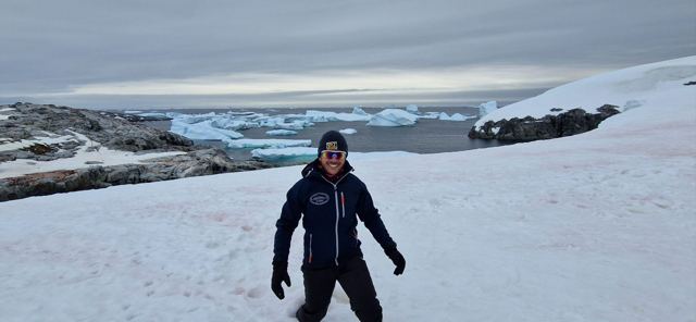 A man in a dark jacket standing on a snowy slope above an icy bay.