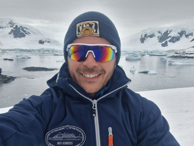 A smiling man in mirrored sunglasses and a beanie with icy mountains behind.