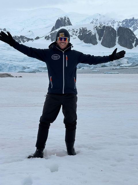 A man with arms outstretched standing in deep snow with ice-covered mountains behind.