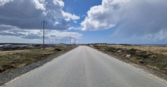 Straight empty road with telegraph poles stretching across flat land under a cloudy blue sky.