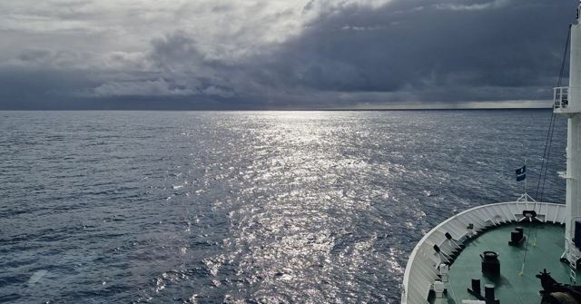 Grey ocean with sunlight glinting under dark clouds, seen from the bow of a ship.
