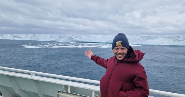 Smiling man in a red coat and beanie on a ship deck gesturing towards a huge iceberg.
