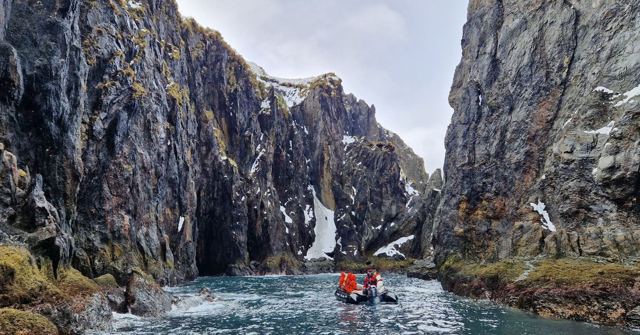Small inflatable boat in a narrow inlet between steep dark cliffs.