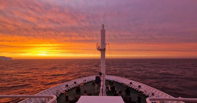 Brilliant orange and pink sky over the sea seen from the bow of a ship.