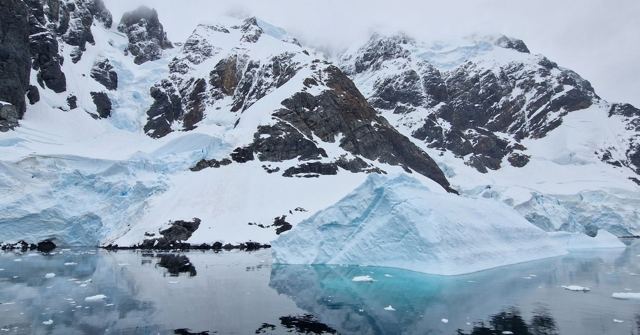 Snow-covered mountains and blue icebergs reflected in calm water.