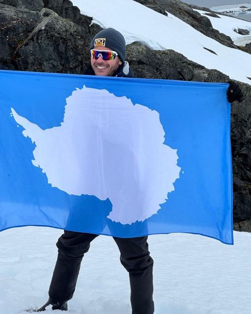 Man in a beanie and sunglasses holding up a blue Antarctica flag on a snowy slope.