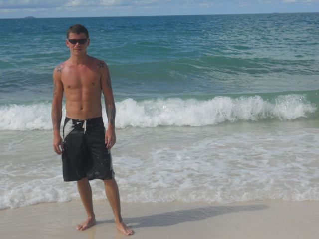 A man in board shorts standing at the water's edge on a beach with waves behind him.