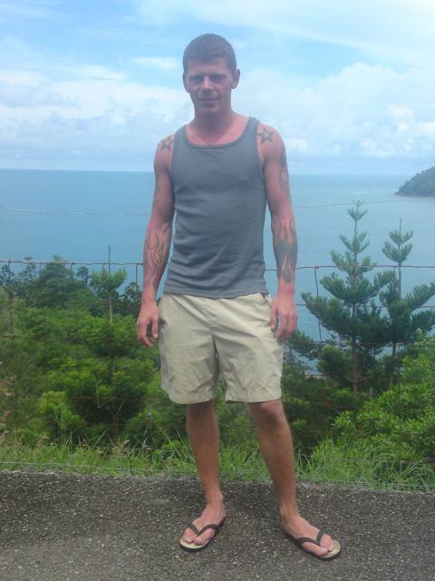 A man in a vest and shorts standing at a lookout above the sea with pine trees behind.