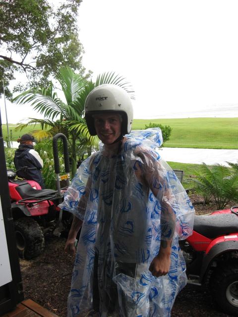 A man in a helmet and clear rain poncho standing by quad bikes under palm trees.