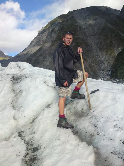 A man in waterproofs holding an ice axe on the white ice of Fox Glacier with mountains behind.