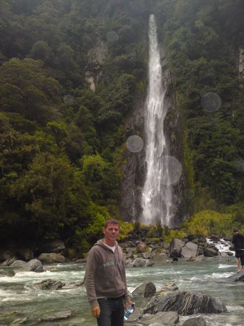A man standing on river rocks below a tall waterfall in a forested gorge.