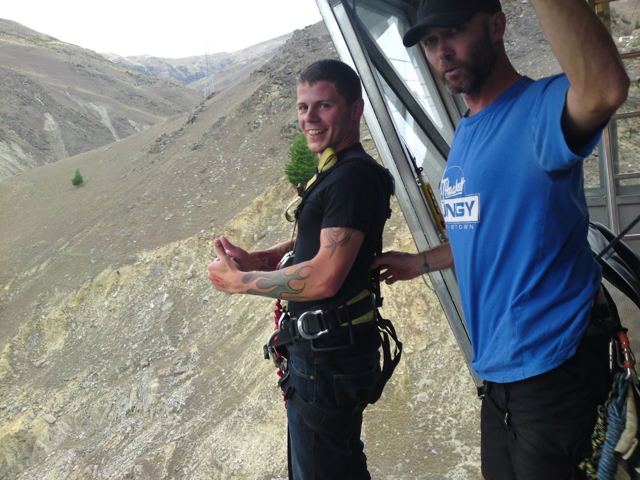 A man in a harness giving a thumbs up in the cable car over a canyon, an instructor beside him.