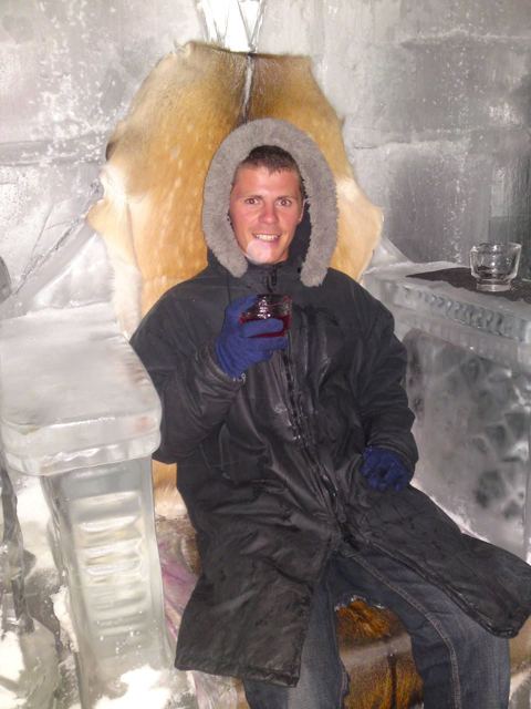 A man in a fur-hooded parka and gloves sitting on a throne carved from ice.