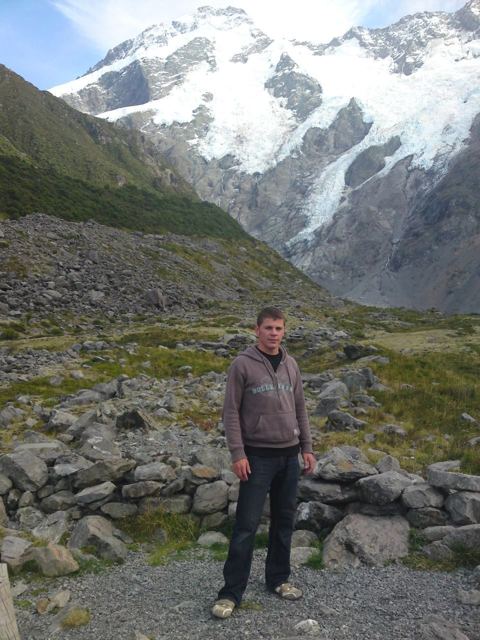 A man standing on a rocky path with snowy mountains and glaciers behind in Mount Cook National Park.