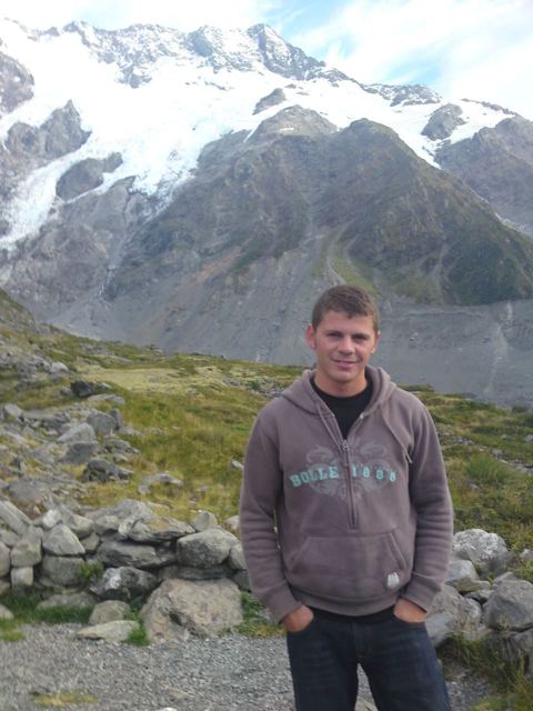 A smiling man in a hoodie with the glaciers and snowy peaks of Aoraki/Mount Cook behind him.
