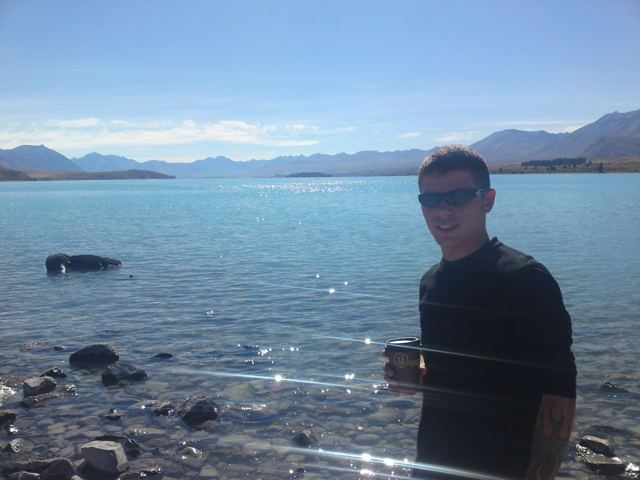 A man in sunglasses standing by a turquoise lake with mountains in the distance.