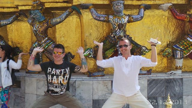 Two men flexing in front of golden demon guardian statues at a temple in Bangkok.