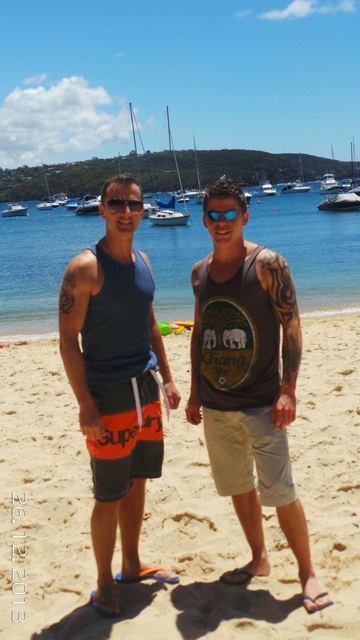 Two men in vests and sunglasses standing on a sandy harbour beach with boats behind.