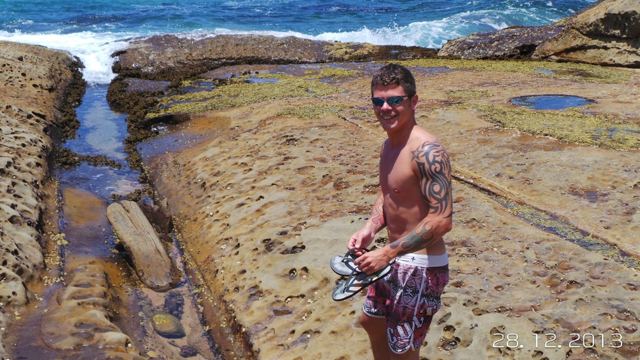 A man in board shorts standing on flat sandstone rock pools by the sea.